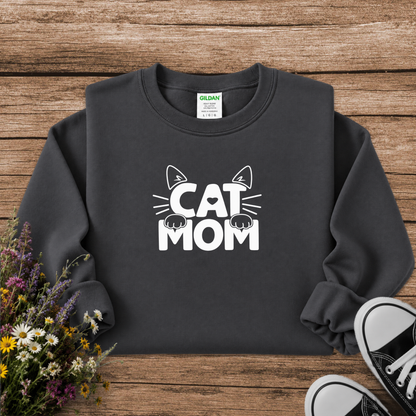 A dark charcoal gray crewneck sweatshirt with a white graphic reading 'CAT MOM' and cat ears/paws, laid flat on a rustic wood background with flowers and black sneakers as props. A Gildan tag is visible inside the collar.