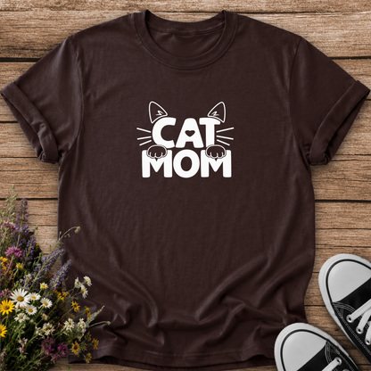A dark brown short-sleeve t-shirt with a white graphic reading 'CAT MOM' and cat ears/paws, laid flat on a wooden surface with flowers and black-and-white sneakers as props.