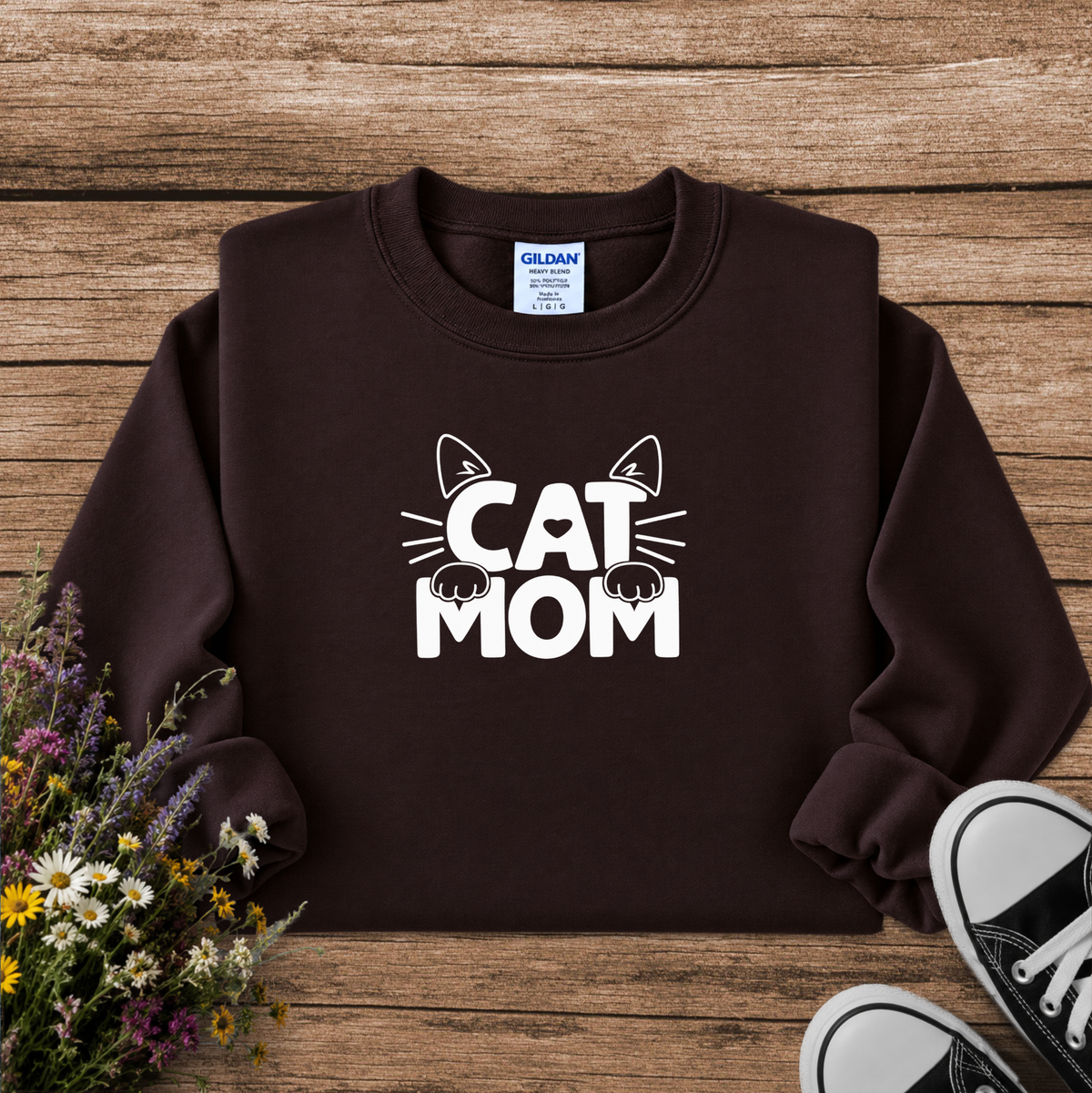 A dark brown crewneck sweatshirt with a white graphic reading 'CAT MOM' and cat ears/paws, laid flat on a rustic wood background with flowers and black sneakers nearby.
