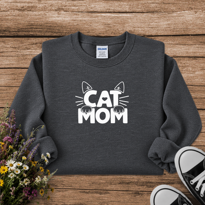 A dark gray crewneck sweatshirt with a white graphic reading 'CAT MOM' and cat ears/paws, laid flat on a rustic wood background with flowers and black sneakers as props.