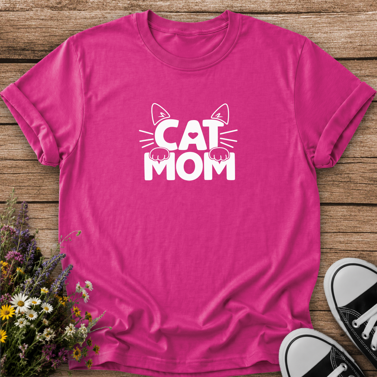 A bright pink short-sleeve t-shirt with a white graphic reading 'CAT MOM' and cat ears/paws, laid flat on a rustic wood surface with flowers and black-and-white sneakers as props.