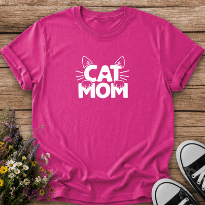 A bright pink short-sleeve t-shirt with a white graphic reading 'CAT MOM' and cat ears/paws, laid flat on a rustic wood surface with flowers and black-and-white sneakers as props.