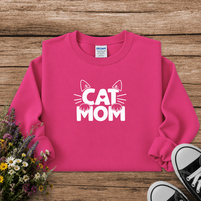 A bright pink crewneck sweatshirt with a white graphic reading 'CAT MOM' and cat ears/paws, laid flat on a rustic wood surface with flowers and black sneakers as props. A Gildan tag is visible inside the collar.