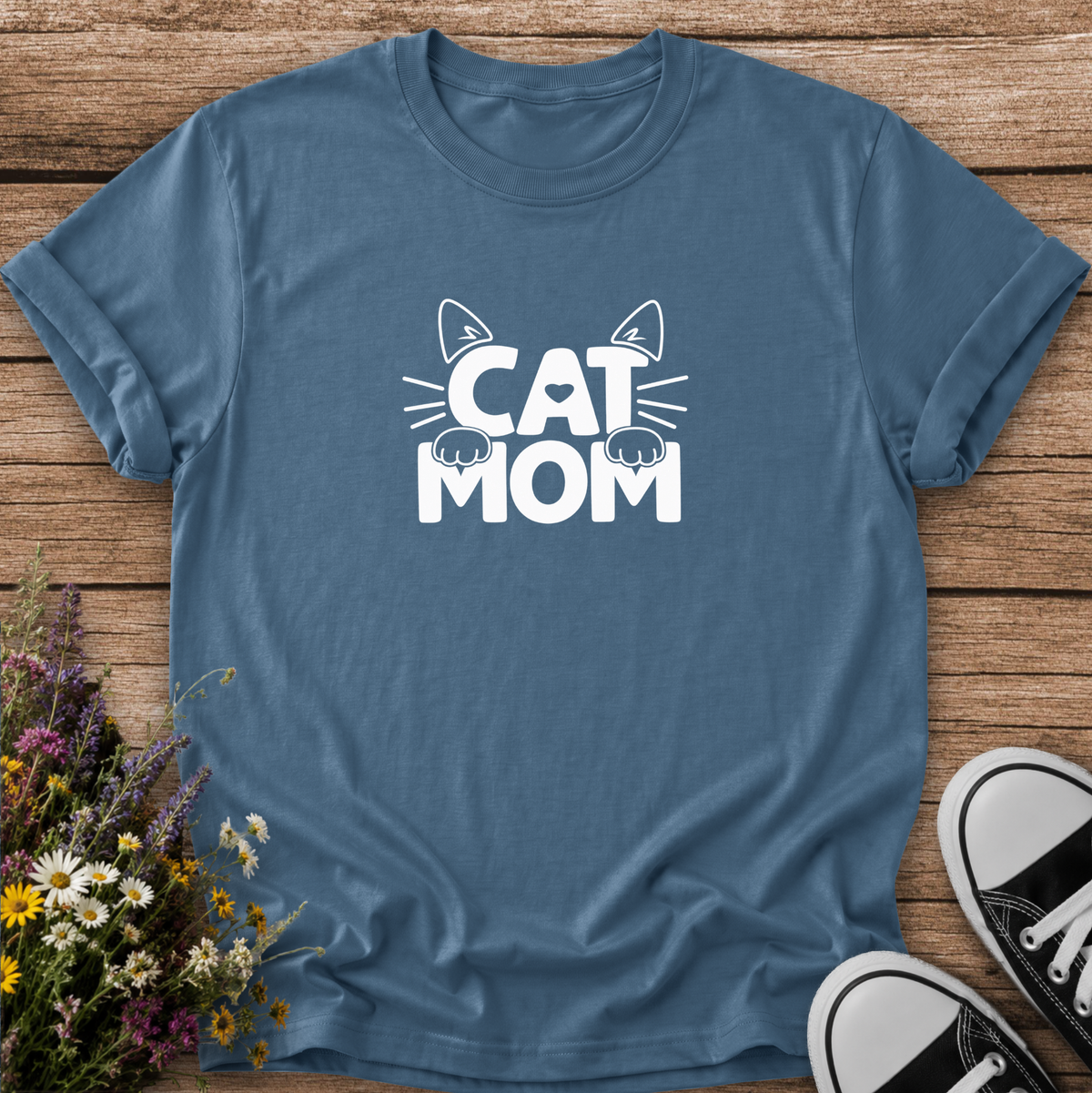 A slate blue crewneck t-shirt with a white 'CAT MOM' graphic featuring cat ears and paws, laid flat on a rustic wooden background with wildflowers and black-and-white sneakers.