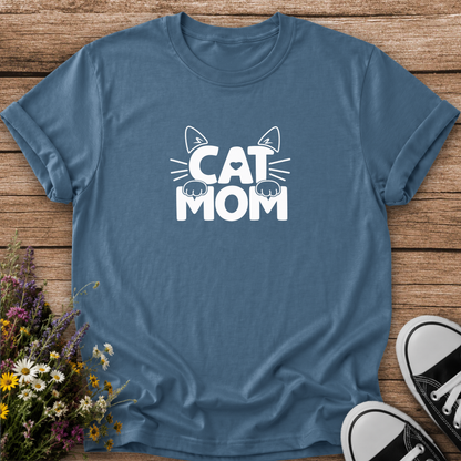 A slate blue crewneck t-shirt with a white 'CAT MOM' graphic featuring cat ears and paws, laid flat on a rustic wooden background with wildflowers and black-and-white sneakers.