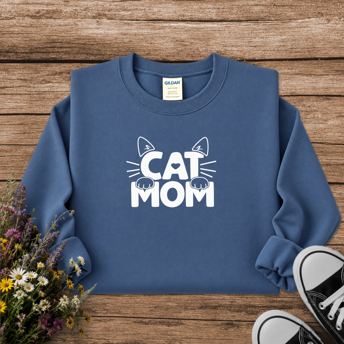 A slate blue crewneck sweatshirt with a white 'CAT MOM' graphic featuring cat ears and paws, laid flat on a rustic wooden background with wildflowers and black sneakers.