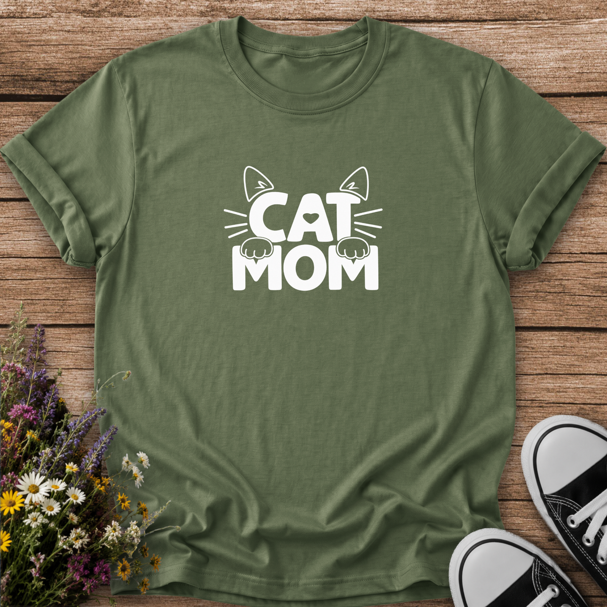 An olive green short-sleeve t-shirt with a white graphic reading 'CAT MOM' and cat ears/paws, laid flat on a rustic wood background with flowers and black-and-white sneakers as props.