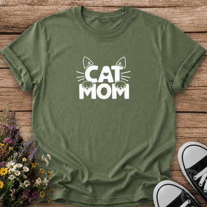 An olive green short-sleeve t-shirt with a white graphic reading 'CAT MOM' and cat ears/paws, laid flat on a rustic wood background with flowers and black-and-white sneakers as props.