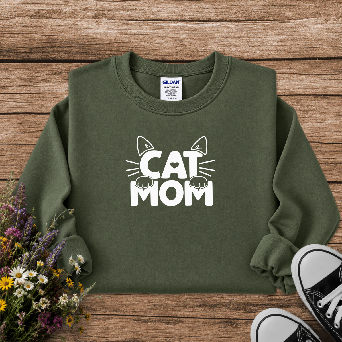 An olive green crewneck sweatshirt with a white graphic reading 'CAT MOM' and cat face details, laid flat on a rustic wood background with flowers and black sneakers as props. A Gildan brand tag is visible inside the collar.