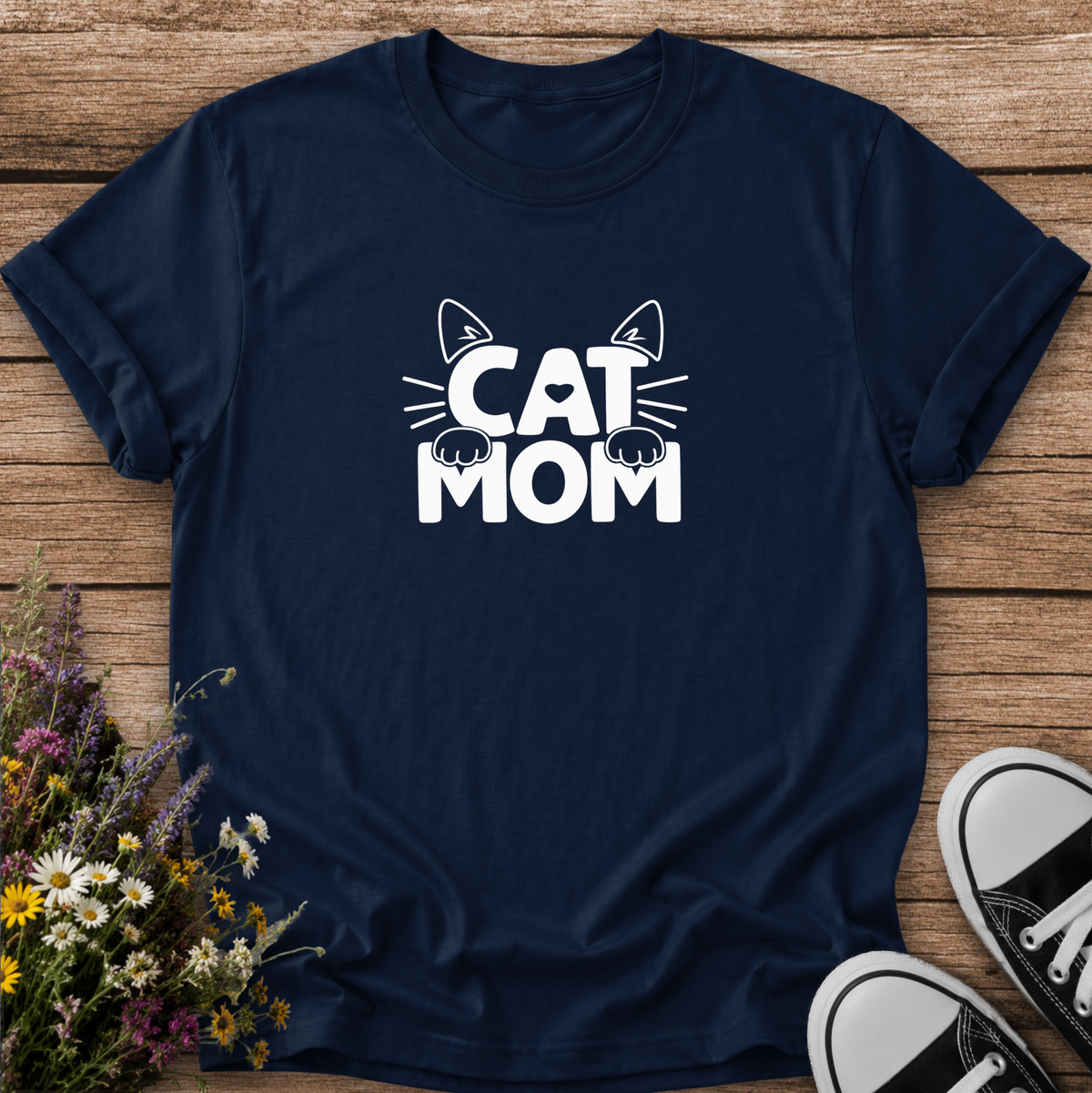 A navy blue short-sleeve t-shirt with a white graphic reading 'CAT MOM' and cat ears/paws, laid flat on a wooden surface with flowers and black-and-white sneakers as props.