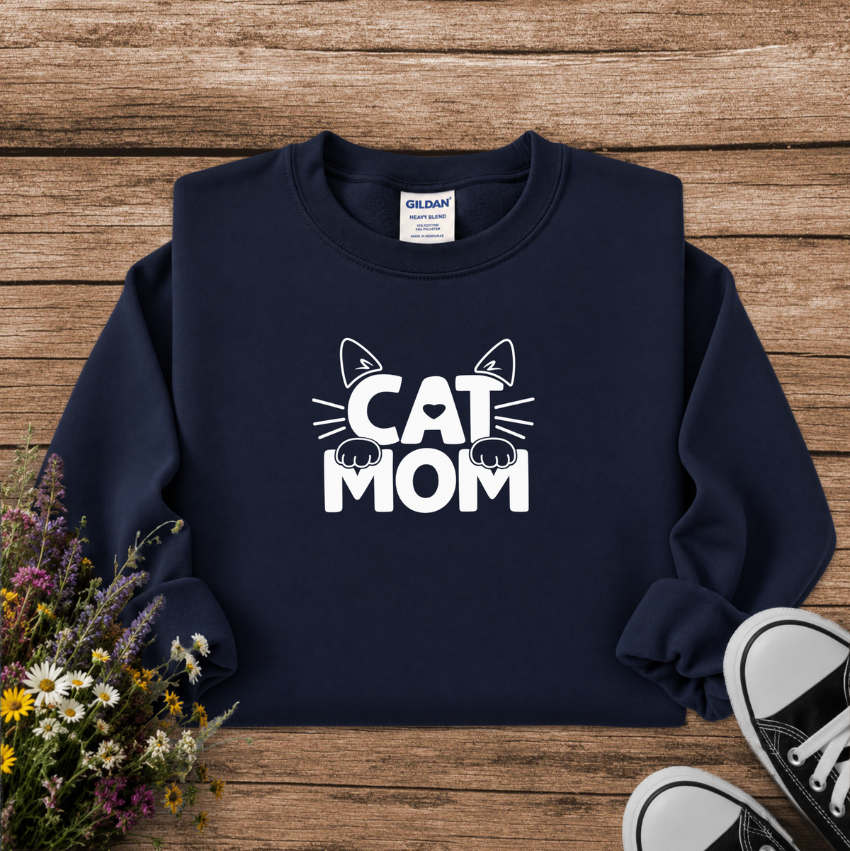 A navy blue crewneck sweatshirt with a white graphic reading 'CAT MOM' and cat ears/eyes, laid flat on a rustic wood background with flowers and black-and-white sneakers as props. Gildan brand label is visible inside the collar.