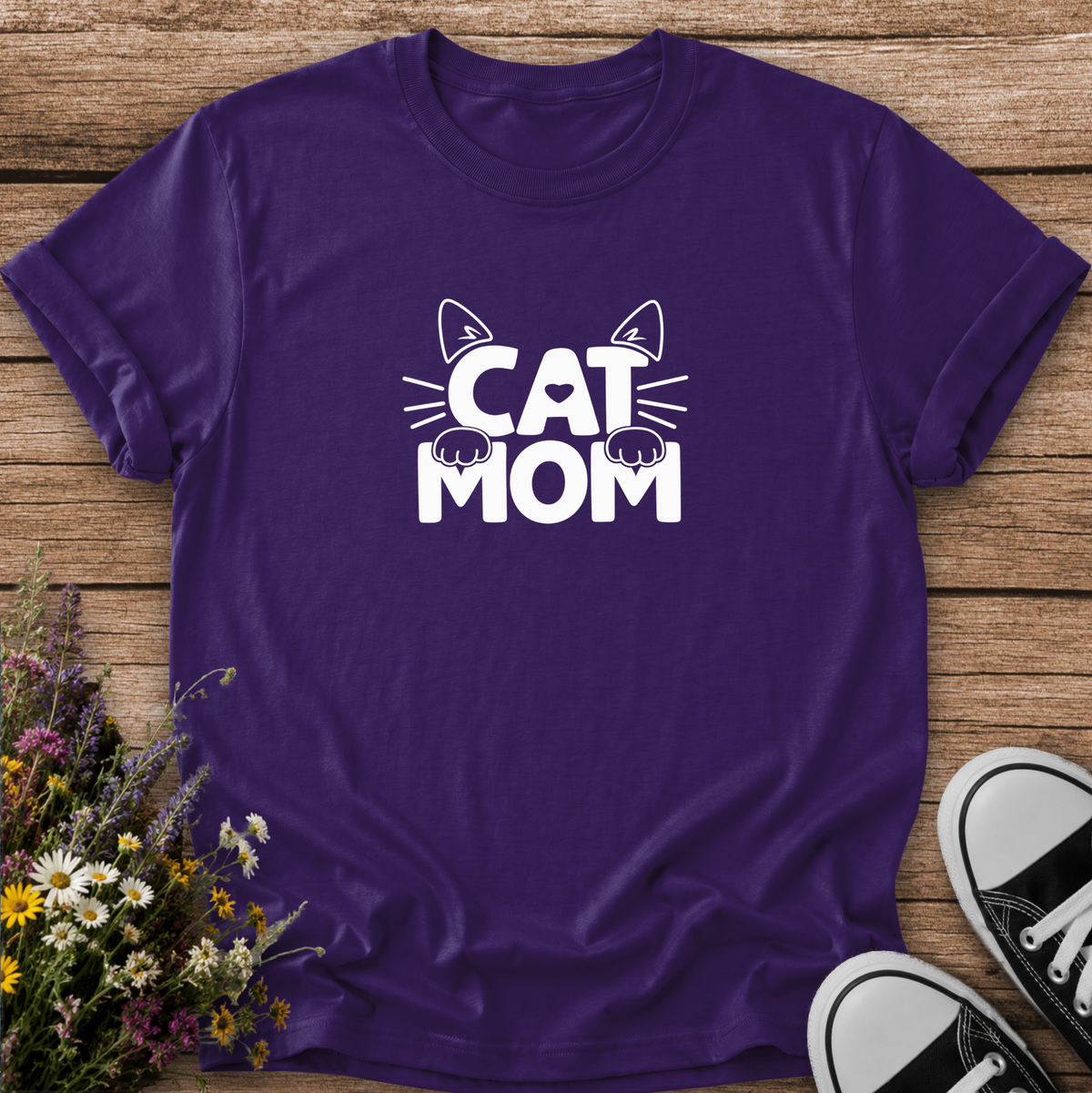 A purple short-sleeve t-shirt with a white 'CAT MOM' graphic featuring cat ears, whiskers, and paws, laid flat on a rustic wooden background with flowers and black-and-white sneakers as props.