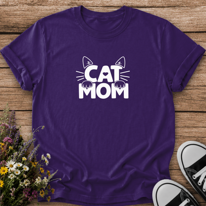 A purple short-sleeve t-shirt with a white 'CAT MOM' graphic featuring cat ears, whiskers, and paws, laid flat on a rustic wooden background with flowers and black-and-white sneakers as props.