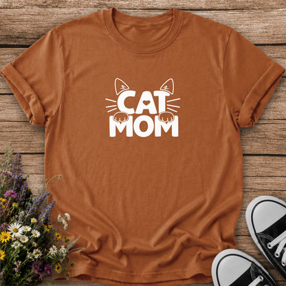 A rust-orange crewneck t-shirt with a white graphic reading 'CAT MOM' with cat ears and paws, laid flat on a rustic wood background with flowers and black-and-white sneakers as props.