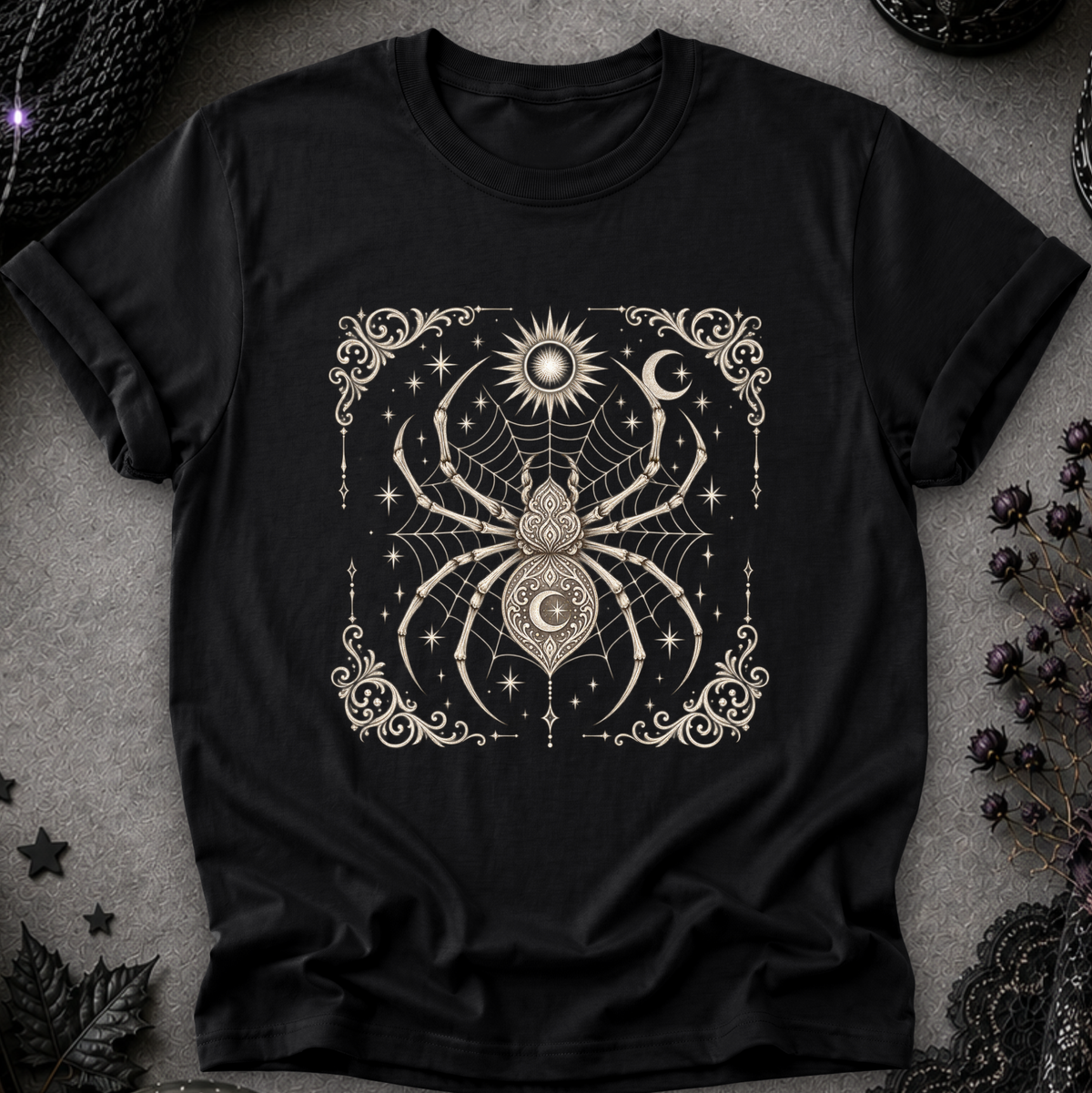 A black short-sleeve t-shirt with a detailed cream-colored gothic graphic of a spider, moon, sun, and stars, laid flat on a textured gray background with dark decorative props.