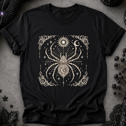 A black short-sleeve t-shirt with a detailed cream-colored gothic graphic of a spider, moon, sun, and stars, laid flat on a textured gray background with dark decorative props.
