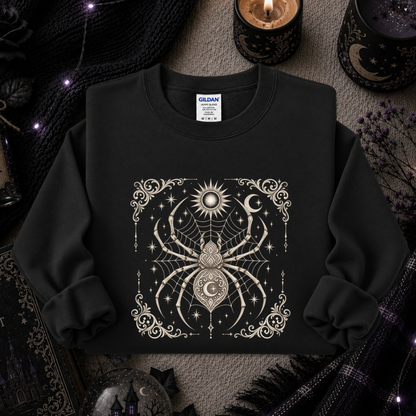 A black crewneck sweatshirt with a detailed cream-colored occult spider graphic on the front, styled in a cozy, dark-themed flat lay with candles, a knit blanket, and occult decor.