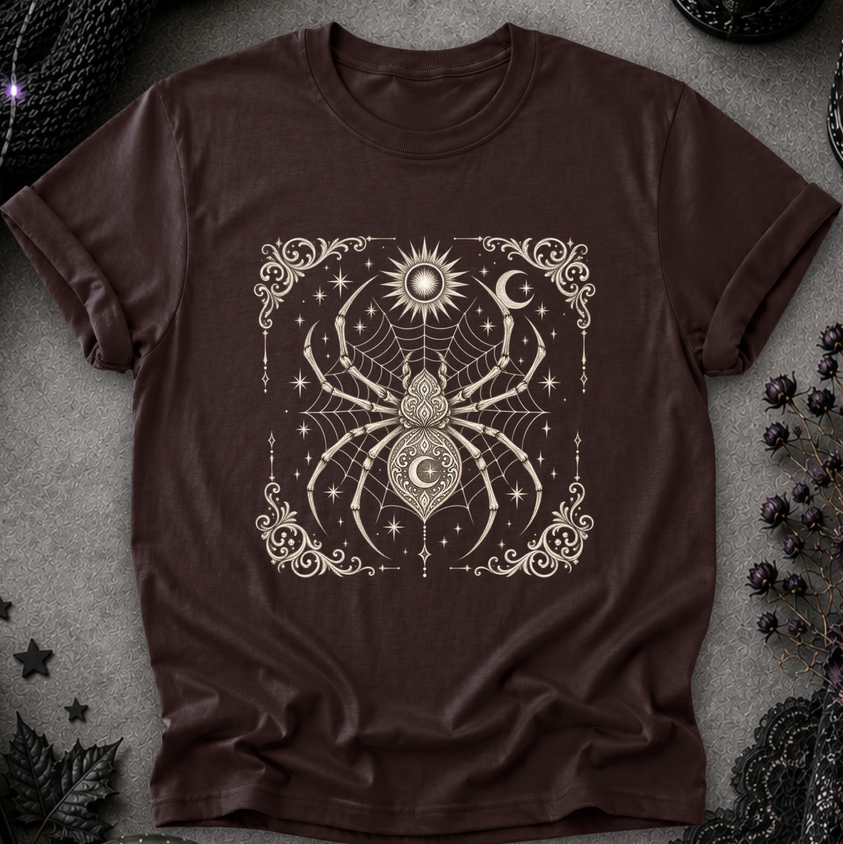 A dark brown short-sleeve t-shirt with a detailed cream-colored occult and celestial graphic featuring a spider, moon, sun, and stars, laid flat on a textured gray background with decorative black lace and dried flowers.