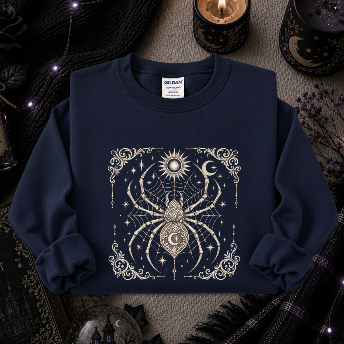 A navy blue crewneck sweatshirt with a detailed cream-colored celestial spider graphic on the front, styled in a cozy, dark-themed flat lay with candles, a knit blanket, and occult decor.