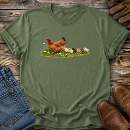 Green t-shirt with a chicken and chicks design on a wooden surface with jeans and boots.