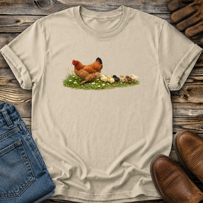Beige t-shirt with a chicken and chicks graphic on a wooden surface with jeans and boots.
