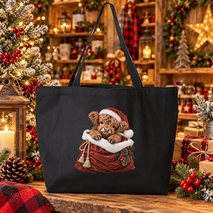 A black canvas tote bag featuring a printed illustration of a fluffy brown highland cow calf in a Santa hat sitting in a red sack, displayed on a rustic wooden table surrounded by a decorated Christmas tree, lanterns, and holiday garlands.