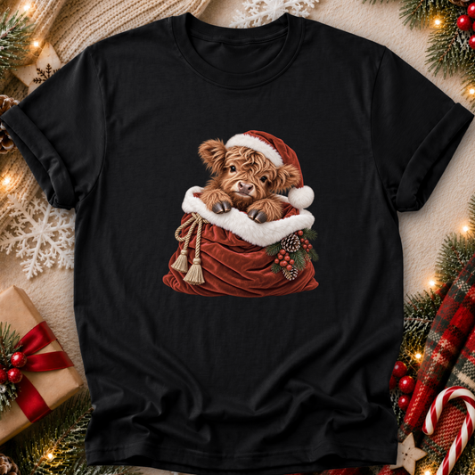 A black short-sleeve t-shirt with a graphic of a fluffy brown Scottish highland cow wearing a Santa hat and sitting in a red Santa sack, surrounded by Christmas decorations like pine branches, lights, and a gift box.