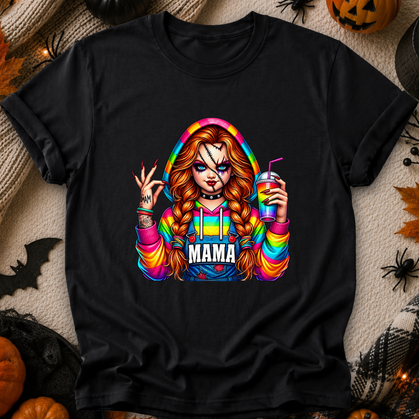 A black short-sleeve graphic T-shirt laid flat on a cozy beige fabric background with Halloween decorations. The shirt features a colorful rainbow-themed graphic of a woman with braided hair holding a drink, with the word 'MAMA' across the front.