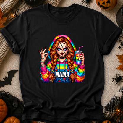 A black short-sleeve graphic T-shirt laid flat on a cozy beige fabric background with Halloween decorations. The shirt features a colorful rainbow-themed graphic of a woman with braided hair holding a drink, with the word 'MAMA' across the front.