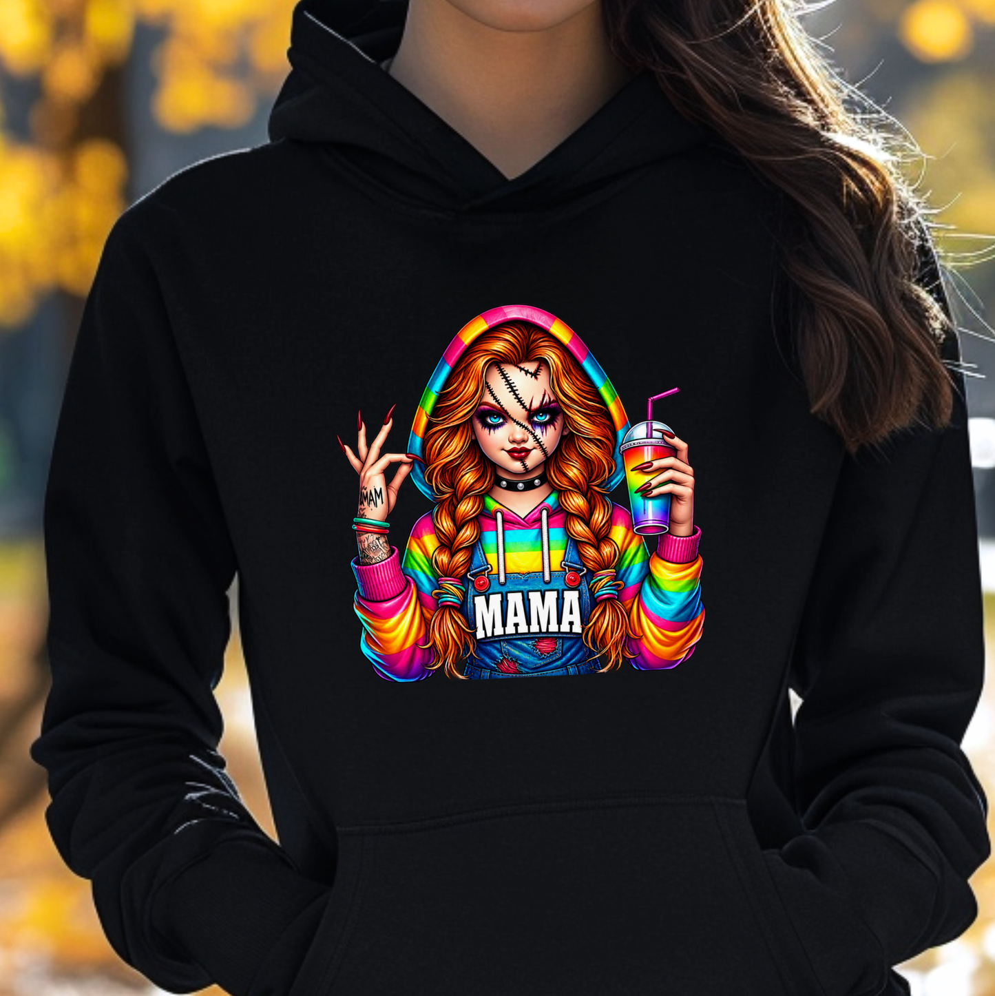A person wearing a black pullover hoodie with a colorful graphic print of a cartoon woman in a rainbow hoodie holding a drink, with the word 'MAMA' printed below. The background is a blurred outdoor scene with yellow autumn foliage.