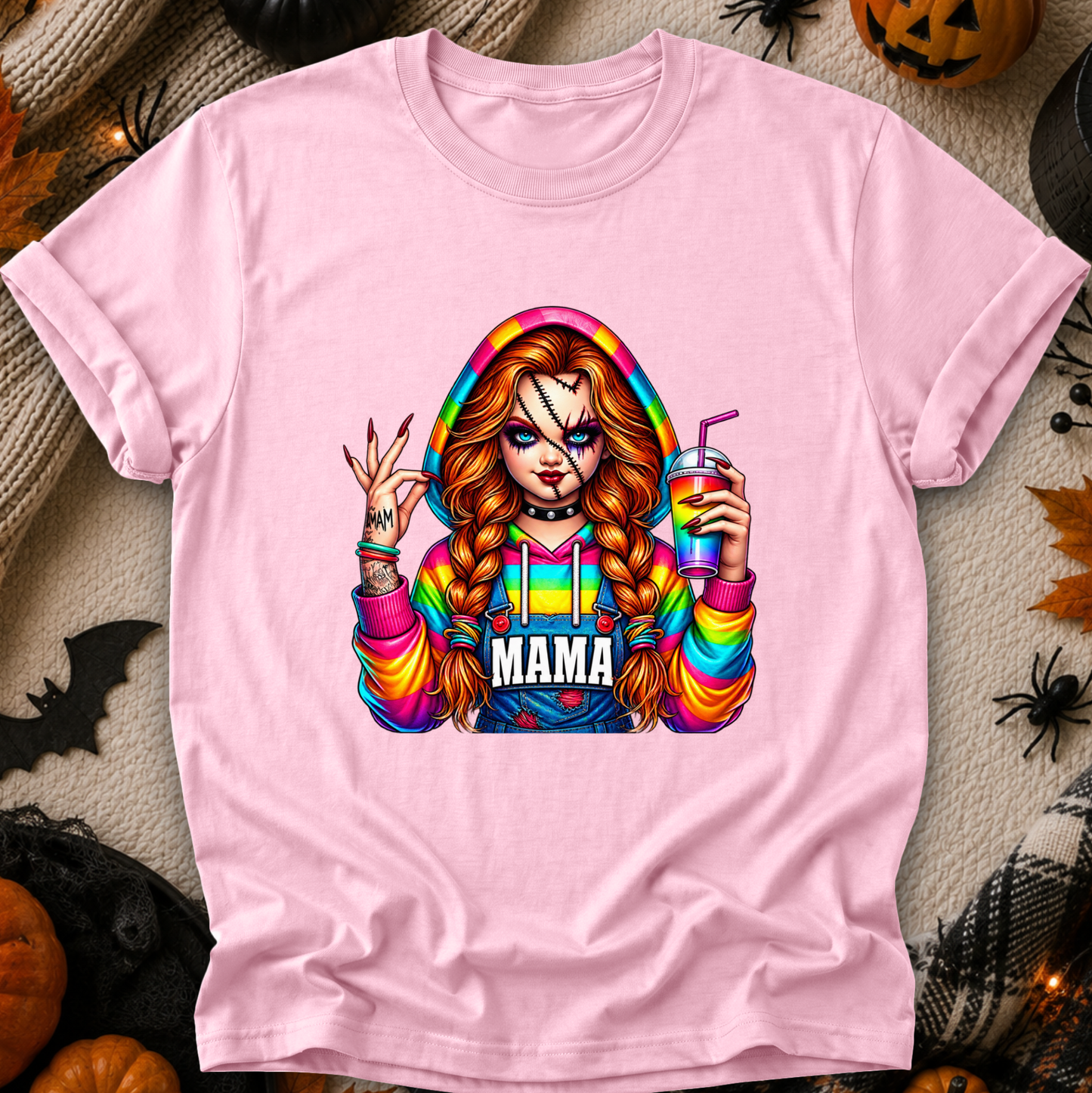 A light pink short-sleeve t-shirt with a colorful graphic of a horror-themed character with the word 'MAMA' on the front, displayed in a flat-lay Halloween setting with pumpkins, spider decorations, and black fabric.