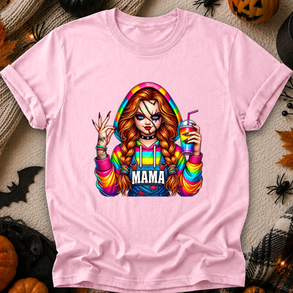 A light pink short-sleeve t-shirt with a colorful graphic of a horror-themed character with the word 'MAMA' on the front, displayed in a flat-lay Halloween setting with pumpkins, spider decorations, and black fabric.