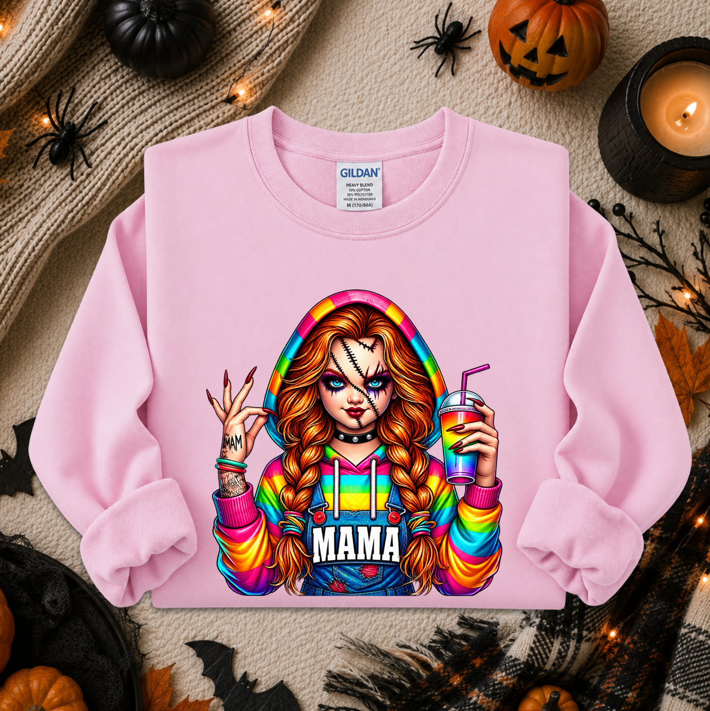 A light pink crewneck sweatshirt by Gildan, featuring a colorful graphic of a woman with long red-blonde braids holding a drink, with the word 'MAMA' printed below. The sweatshirt is displayed in a flat-lay arrangement with Halloween decorations including a pumpkin, spider, candles, and a knit sweater.