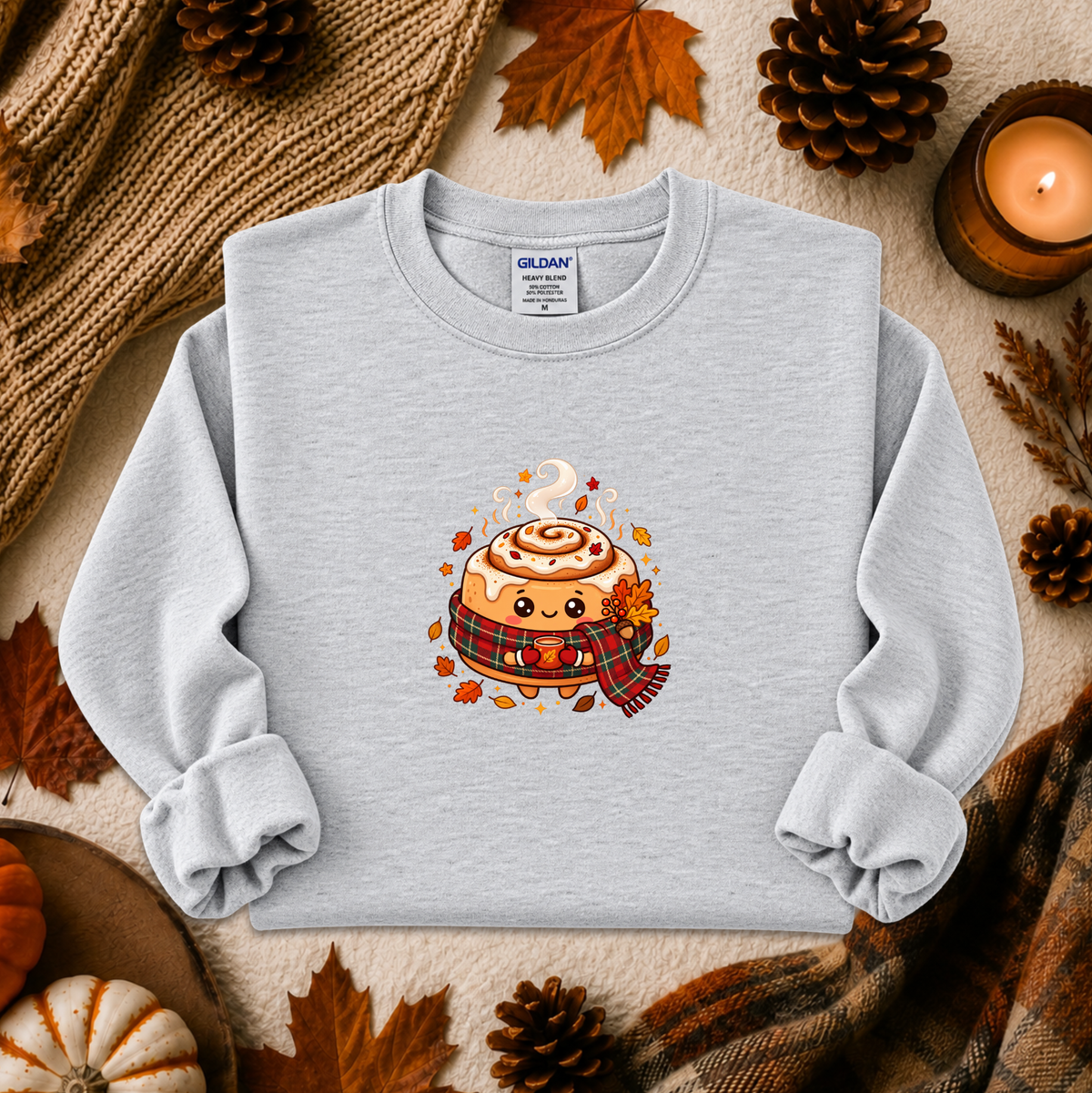 A light gray long-sleeve crewneck sweatshirt with a cute cartoon cinnamon roll graphic on the chest, styled in a flat lay with autumn props including pinecones, leaves, a candle, and a knit blanket.
