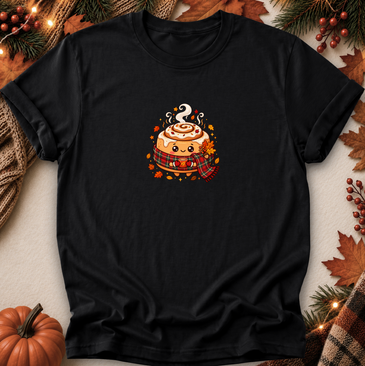 A black short-sleeve crewneck t-shirt laid flat on a neutral background, decorated with autumn props like a pumpkin, pine branches, and leaves. The center of the shirt features a cute cartoon graphic of a cinnamon roll wearing a red plaid scarf and holding a hot cocoa cup.