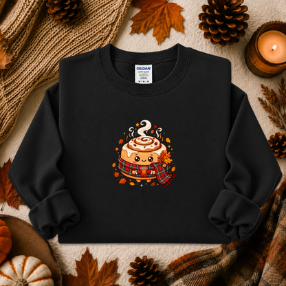 A black crewneck sweatshirt featuring a cute cartoon cinnamon roll graphic with autumn accents, styled in a flat lay with fall leaves, pinecones, a candle, and a cozy knit blanket. The Gildan brand tag is visible inside the collar.
