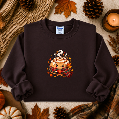 A dark brown crewneck sweatshirt featuring a cute cartoon cinnamon roll graphic with autumn leaves and a plaid scarf, styled in a cozy fall flat-lay with pinecones, a candle, and a knit blanket.