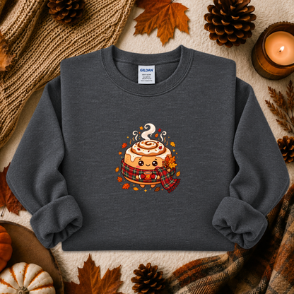 A dark gray crewneck sweatshirt featuring a cute cartoon cinnamon roll graphic with autumn accents, styled in a flat lay with fall-themed props like pinecones, a candle, and a knit blanket.