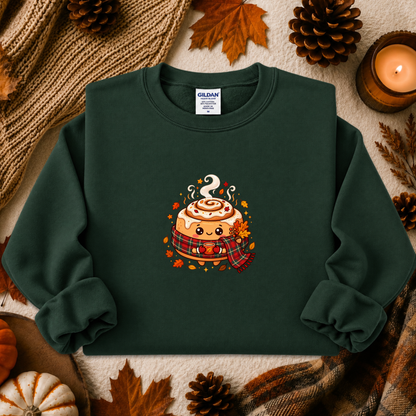 A dark green crewneck sweatshirt with a cute cartoon cinnamon roll graphic, styled in an autumn flat-lay with pinecones, a candle, and a knit blanket.