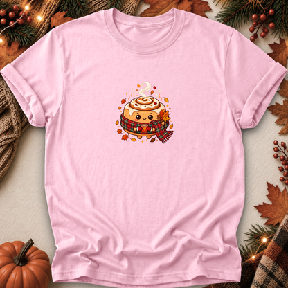 A light pink short-sleeve crewneck t-shirt featuring a cute cartoon cinnamon roll wearing a red plaid scarf, styled in a flat-lay with autumn decorations including a pumpkin, pine branches, and leaves.
