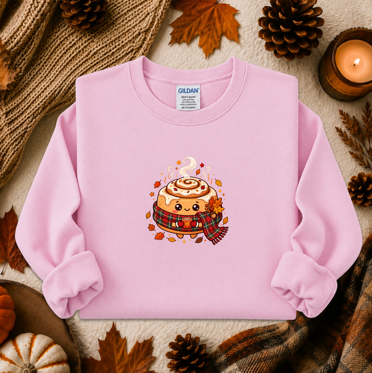 A light pink long-sleeve sweatshirt laid flat on a cozy autumn-themed background with pinecones, leaves, a candle, and a knit blanket. The sweatshirt features a cute cartoon cinnamon roll graphic on the chest with a plaid scarf and fall leaves.