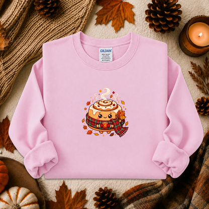 A light pink long-sleeve sweatshirt laid flat on a cozy autumn-themed background with pinecones, leaves, a candle, and a knit blanket. The sweatshirt features a cute cartoon cinnamon roll graphic on the chest with a plaid scarf and fall leaves.