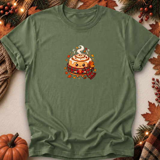 An olive green short-sleeve t-shirt with a cute cartoon cinnamon roll graphic on the chest, styled in a flat-lay with autumn decorations including a pumpkin, pine branches, and leaves.