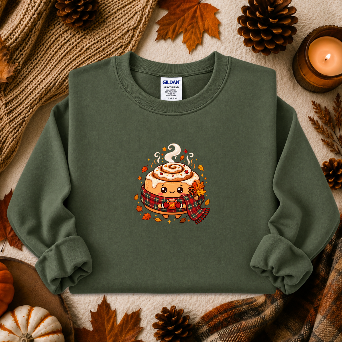An olive green crewneck sweatshirt by Gildan, featuring a cute cartoon cinnamon roll graphic with autumn details, styled in a cozy flat-lay with fall leaves, pinecones, a candle, and a knit blanket.