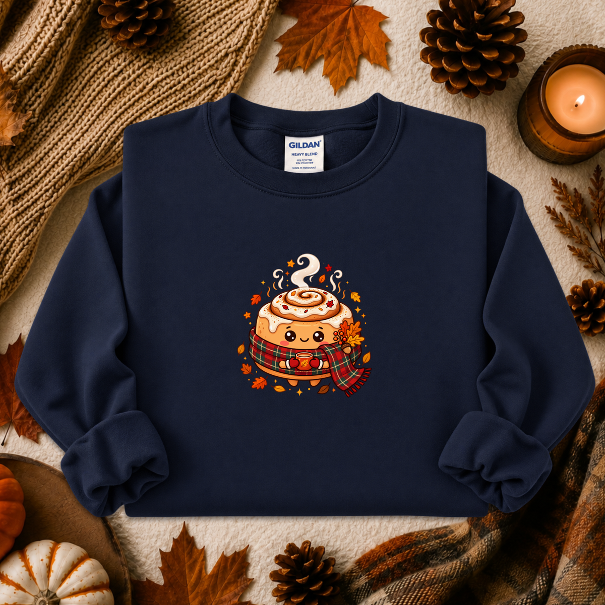 A navy blue crewneck sweatshirt with a cute cartoon cinnamon roll graphic, styled in a cozy autumn flat-lay with pinecones, a candle, and a knit blanket. The brand Gildan is visible on the inner neck label.