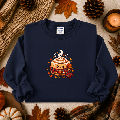 A navy blue crewneck sweatshirt with a cute cartoon cinnamon roll graphic, styled in a cozy autumn flat-lay with pinecones, a candle, and a knit blanket. The brand Gildan is visible on the inner neck label.