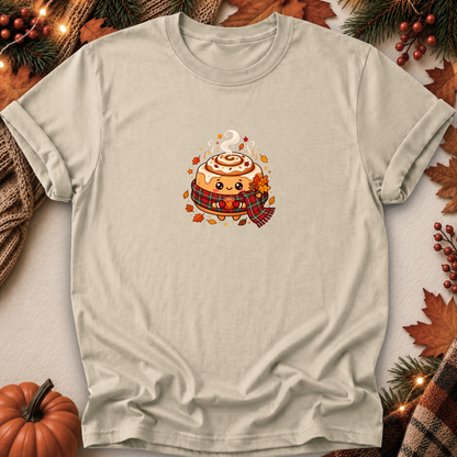 A light beige crewneck t-shirt featuring a cute cartoon cinnamon roll wearing a red plaid scarf, surrounded by autumn leaves and fall decorations like a small pumpkin, pine branches, and string lights.