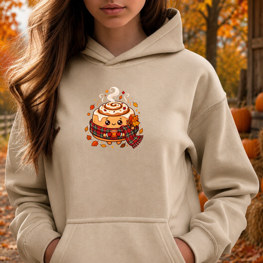 A woman wearing a beige hoodie with a cute cartoon cinnamon roll graphic on the chest, set against an autumn outdoor background with pumpkins and fall foliage.