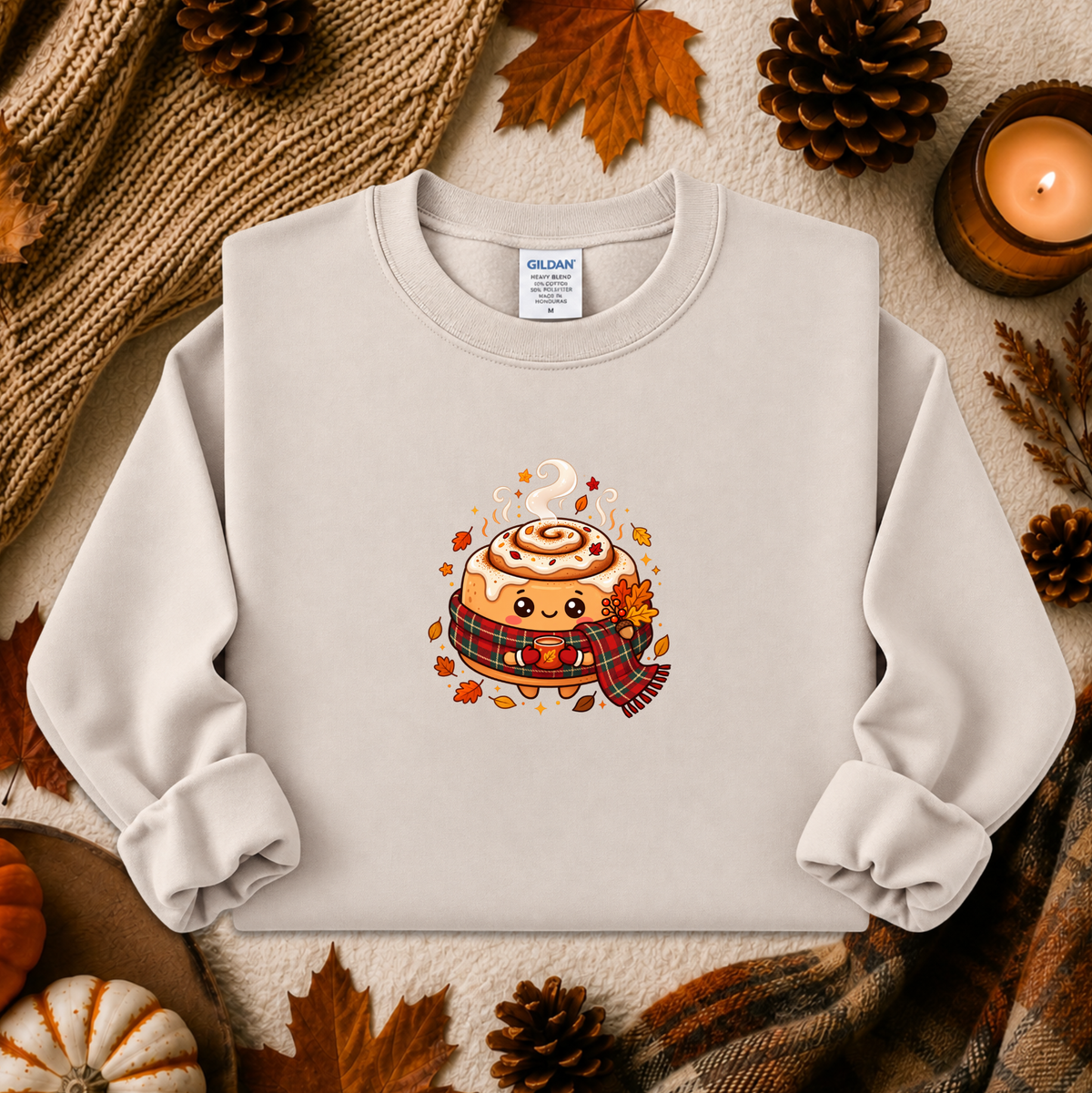 A light gray sweatshirt laid flat with autumn-themed props including pinecones, leaves, a candle, and a knitted scarf. The sweatshirt has a centered graphic of a cute cinnamon roll character wearing a plaid scarf.