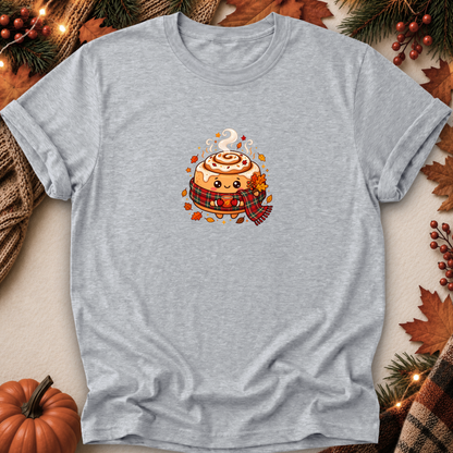 A light heather gray short-sleeve t-shirt with a cute cartoon cinnamon roll graphic on the chest, styled in a flat-lay with autumn decorations like a pumpkin, pine branches, and leaves.
